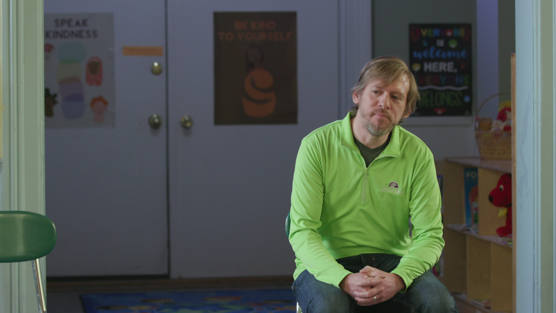 Robert McPherson sits and speaks with cubbies and artwork on a wall and double doors in the background.