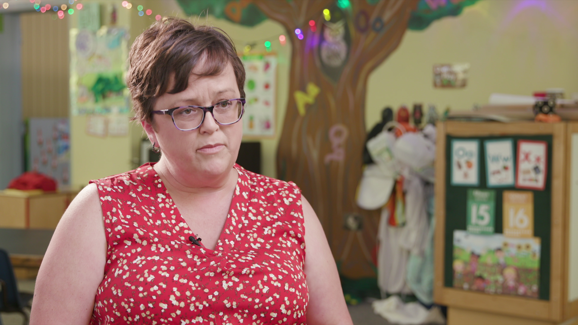 Heather Murray sits and speaks with string lights, artwork and a painted tree on a wall in the background.