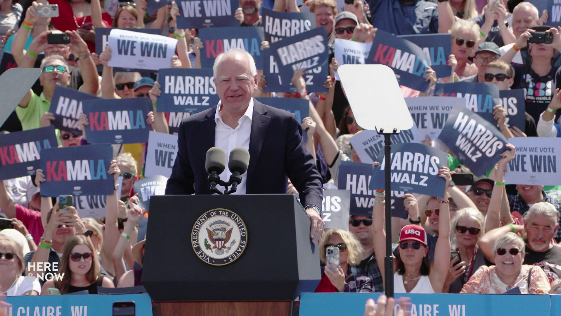 Tim Walz stands and speaks behind a podium with microphones and teleprompter mirrors on either side and people holding signs reading 'Kamala,' 'Harris Walz' 'When We Vote We Win' standing in the background.