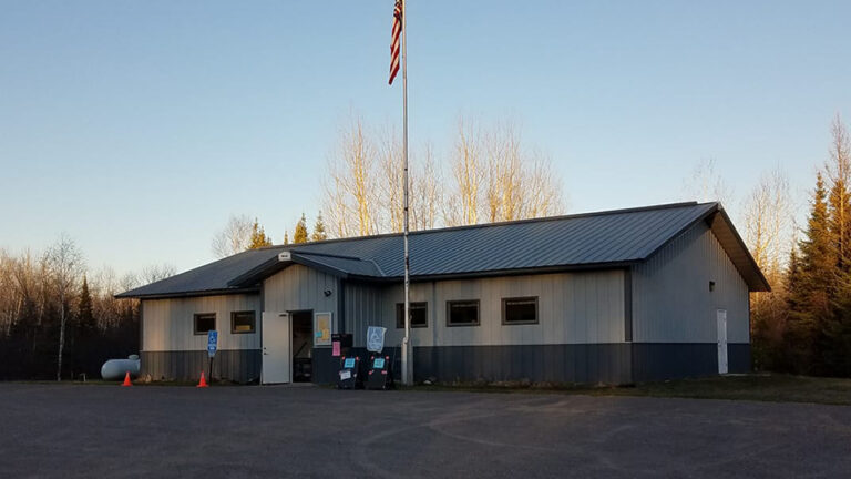 A U.S. flag flies from the top of a flagpole in front of a single-story building with an open door and several windows along its front wall, a propane tank on one side, a parking lot in front, and trees in the background.