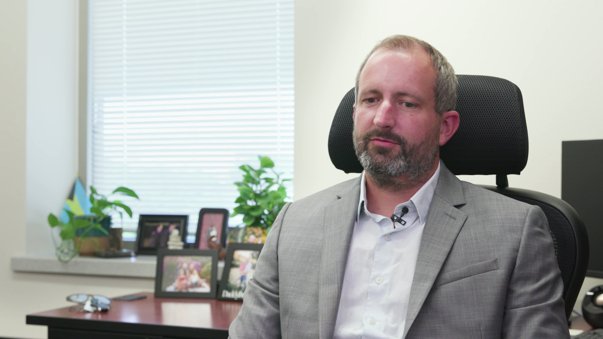 David Helpap speaks while sitting in a room with framed photos and potted plants on a desk and windowsill in the background.