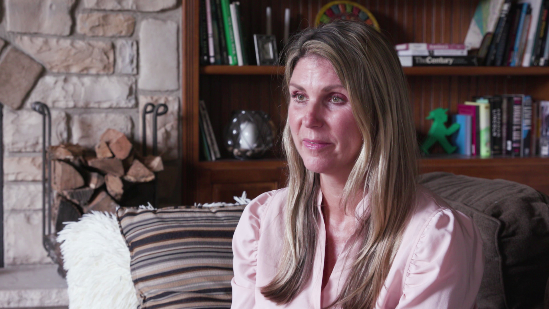 Kristin Lyerly sits while speaking in a room with a wood bookshelf lined with books and other items as well as a stone fireplace with a stack of split logs in the background.