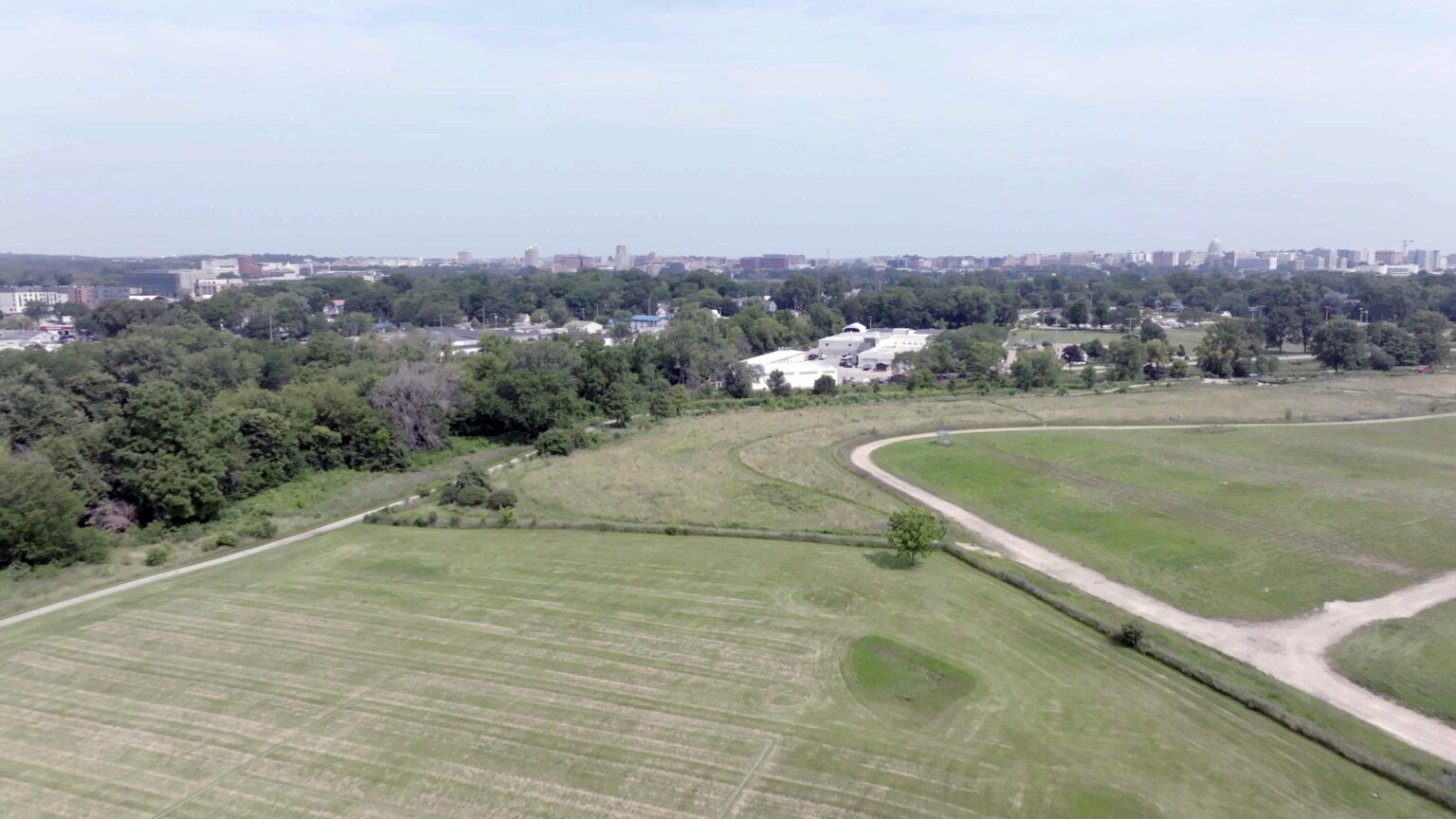 An aerial photos shows gravel paths running though multiple fields of grass, with buildings and trees in the background and an urban area on the horizon with a dome marking the highest reaching building.