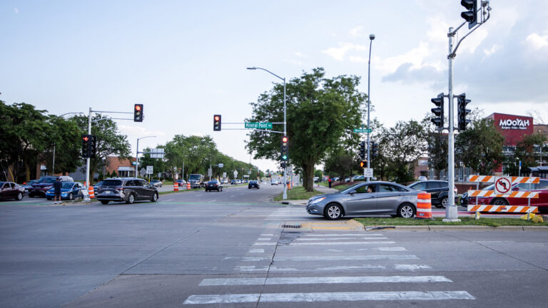 Vehicles drive through and wait at traffic signals at an intersection of two divided roads with crosswalks, medians and multiple lanes, with trees, streetlights and buildings in the background.