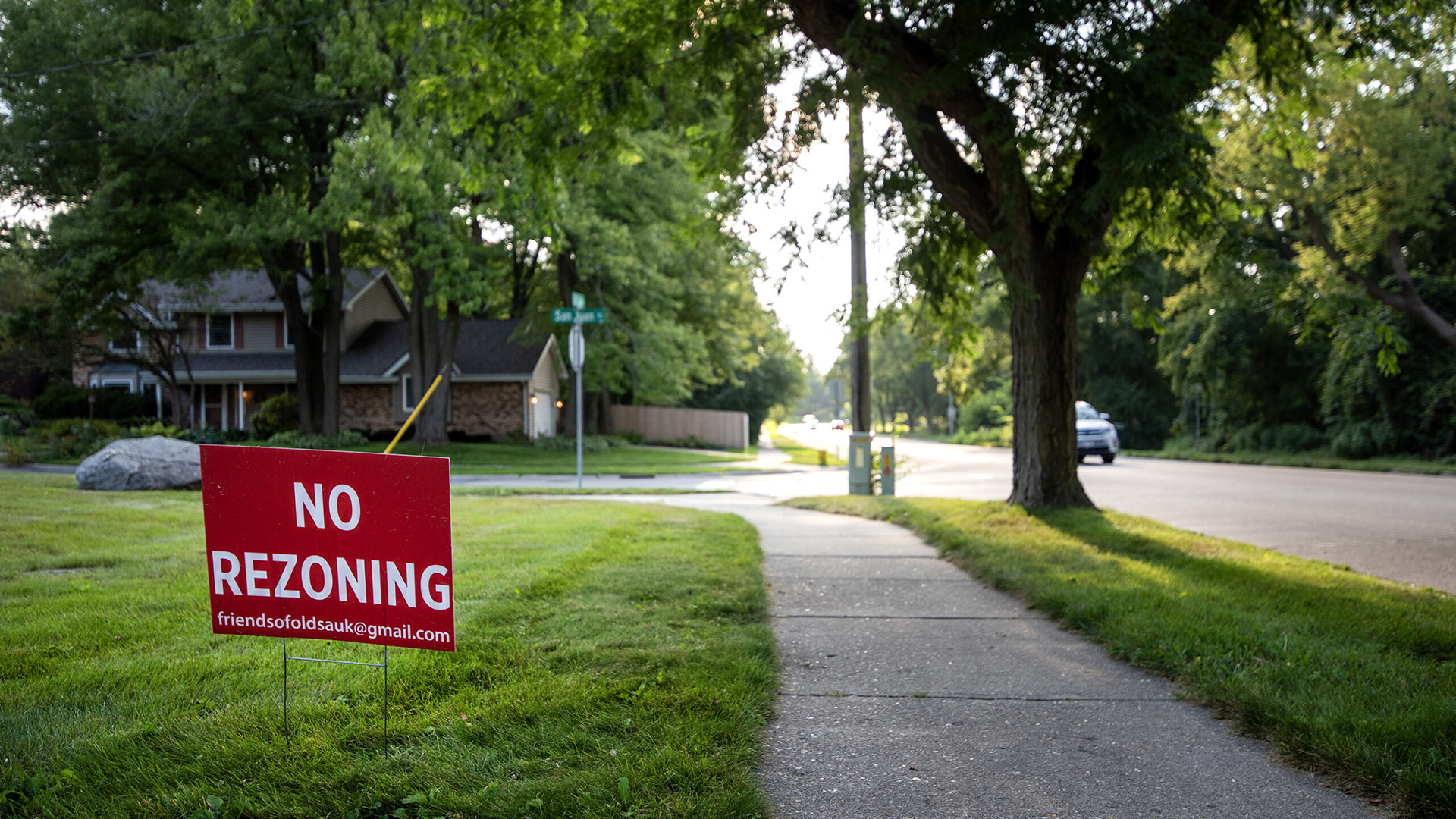 A political yard sign with the words "No Rezoning" and an email address stands in a lawn next to a sidewalk alongside a road with a tree in the terrace, with a house and trees in the background.