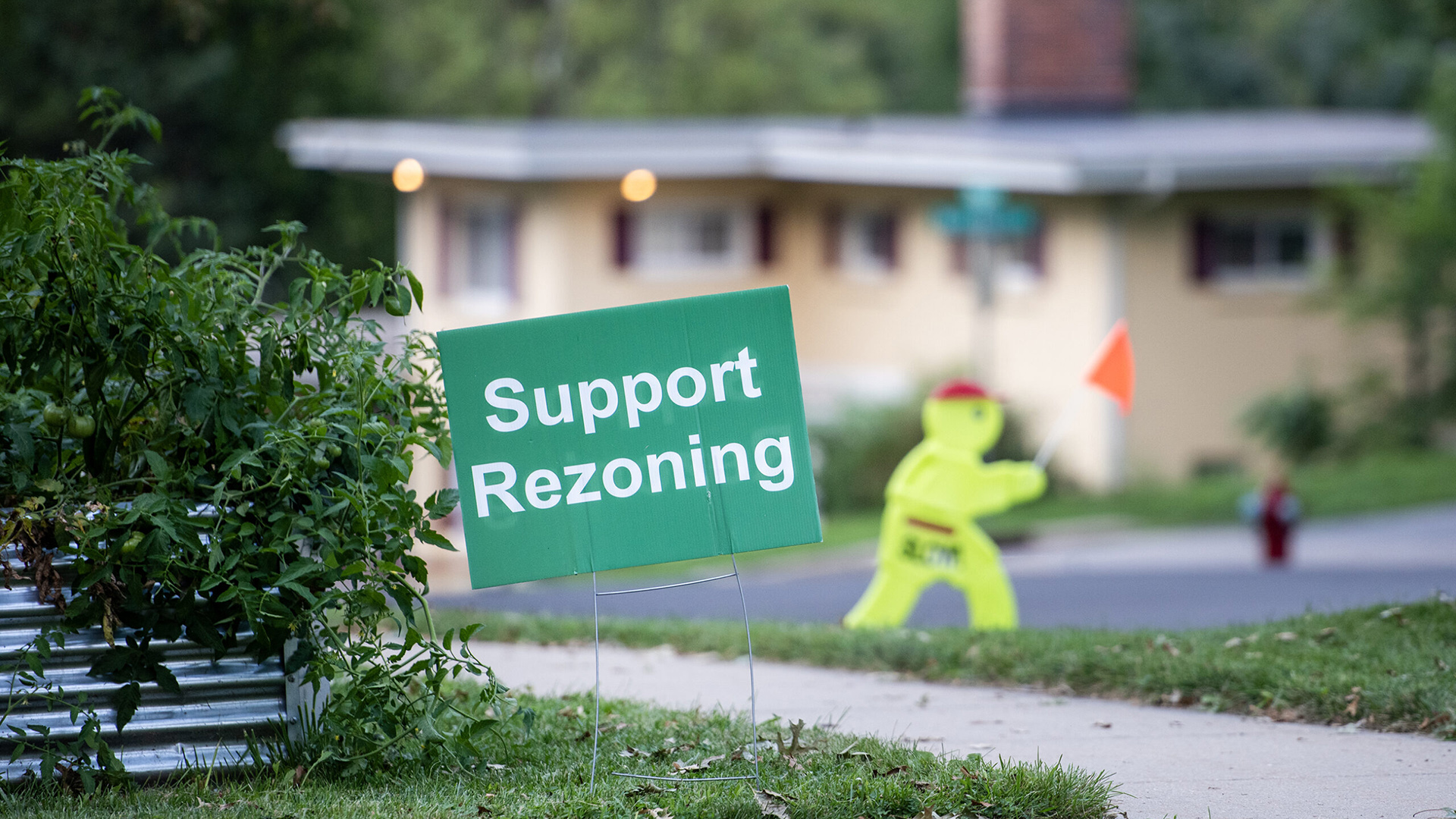 A political yard sign with the words "Support Rezoning" stands in a lawn between a metal planter and a sidewalk alongside a road, with an out-of-focus safety road sign in the form of a walking figure and house in the background.