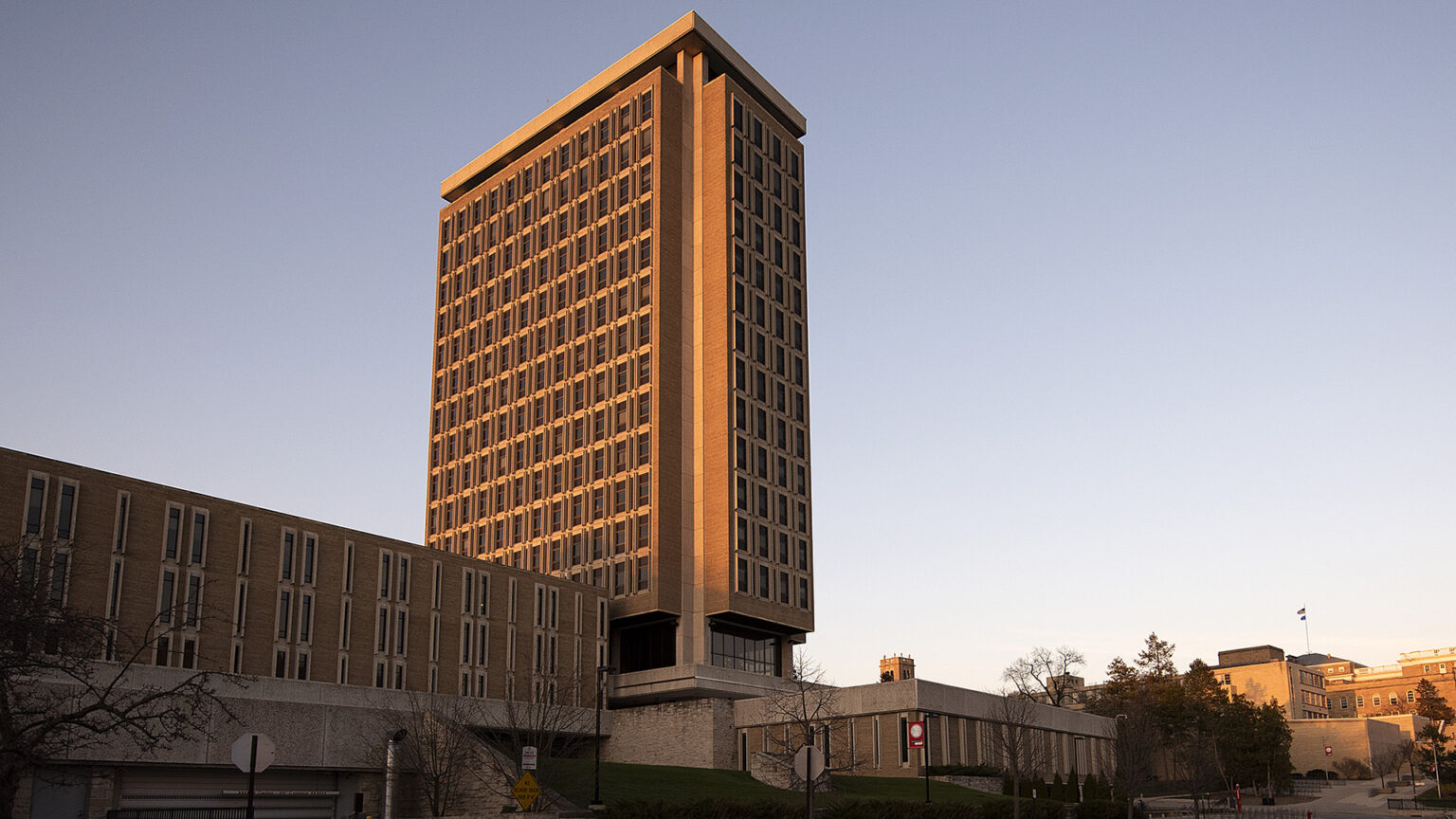 Sunlight illuminates one face of a tall, multi-story portion of a building stands next to multiple, shorter wings, with trees and other buildings in the background under a clear sky.