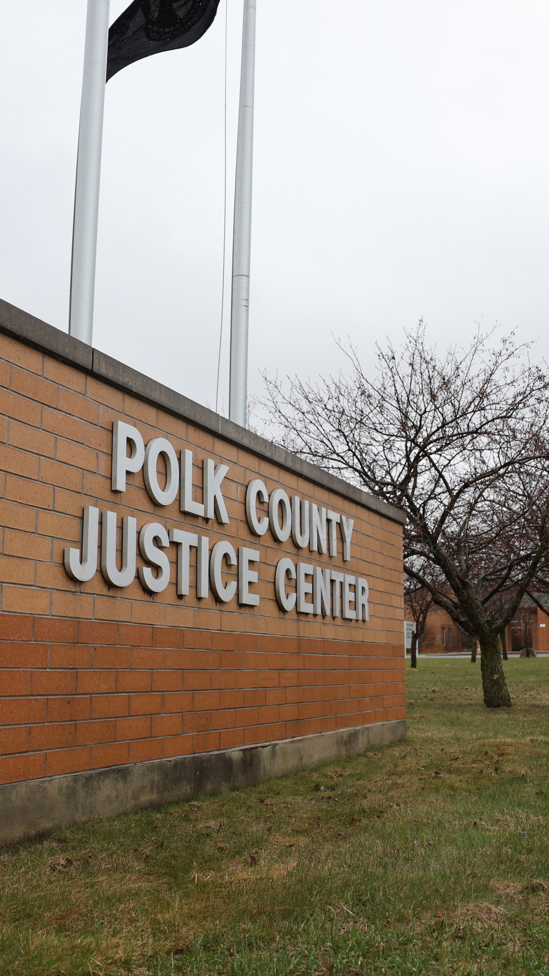 A short brick wall with a letter sign reading "Polk County Justice Center" stands on a lawn next to flagpoles and in front of a row of leafless trees in front of a building.