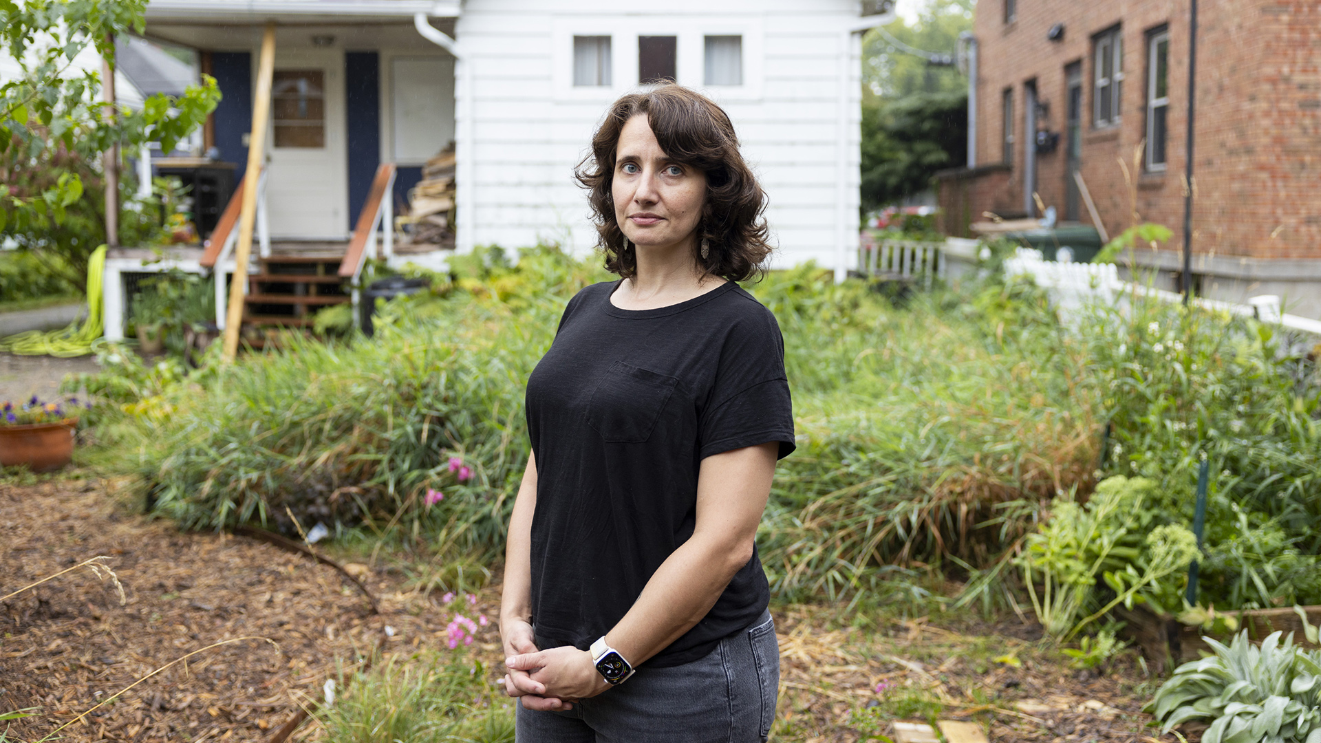 Miriam Hasan poses for a portrait while standing in a backyard with a garden and mulched area and a wood-sided house with a covered porch in the background next to an adjacent brick building with multiple doors and windows.