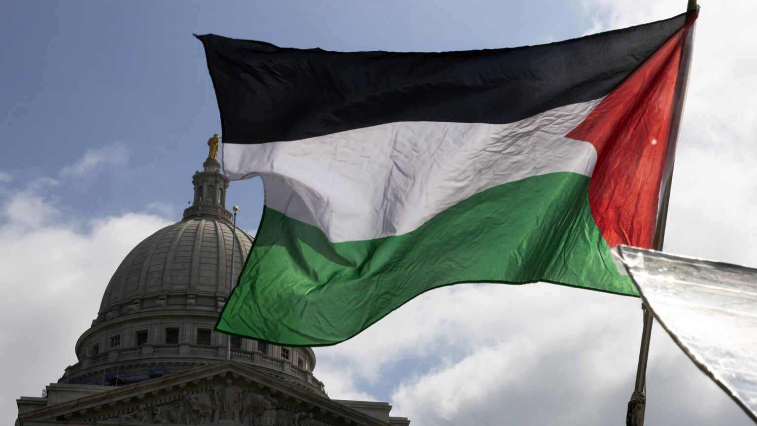 The flag of Palestine flies from a handheld pole, with the dome of the Wisconsin State Capitol in the background under a partly cloudy sky.