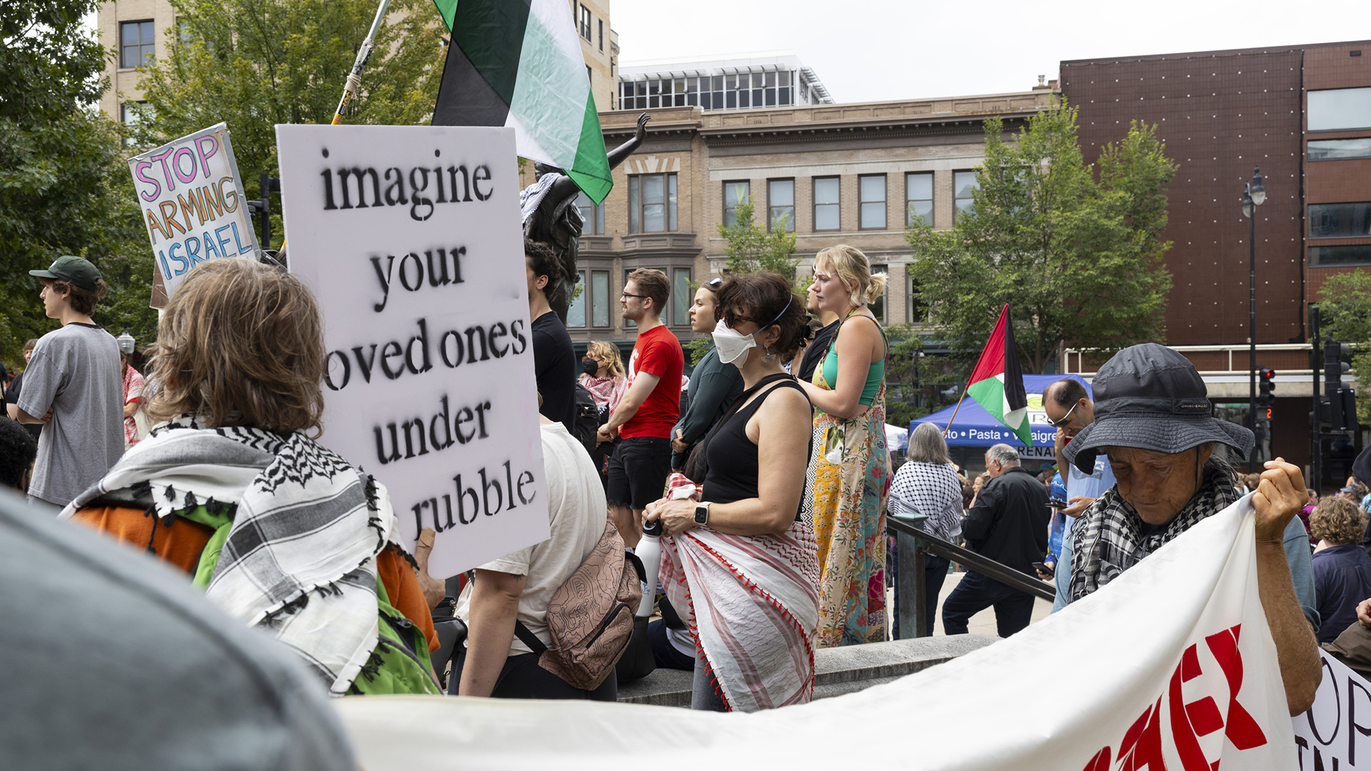 Miriam Hasan stands among a group of people standing on stairs and facing in the same direction, with one holding a sign reading "Stop Arming Israel" and another sign reading "imagine your loved ones under rubble" while other people hold flags of Palestine on handheld poles, with trees and a block of multi-story buildings in the background.