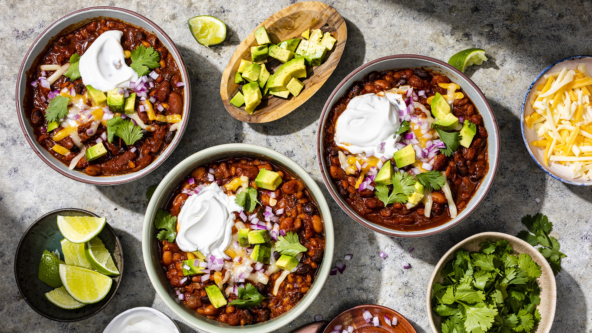 Three large bowls of chili topped with sour cream and avocados sit on a countertop surrounded by bowls of limes, herbs and other toppings.