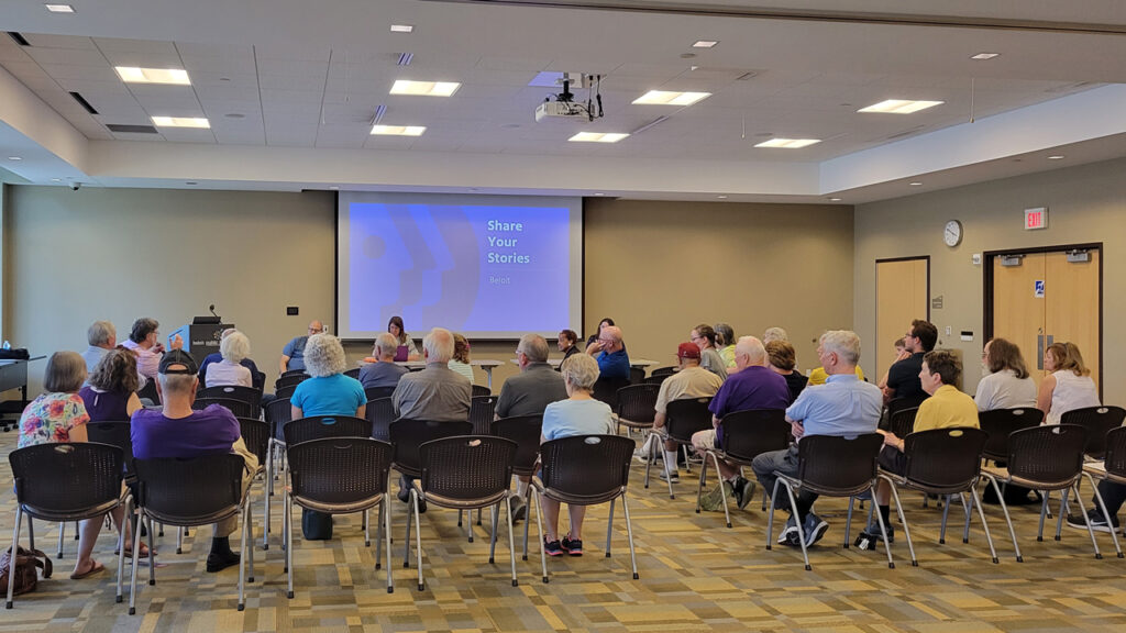 A crowd sitting in a public library to watch a presentation on a projector, "Share your stories, Beloit."