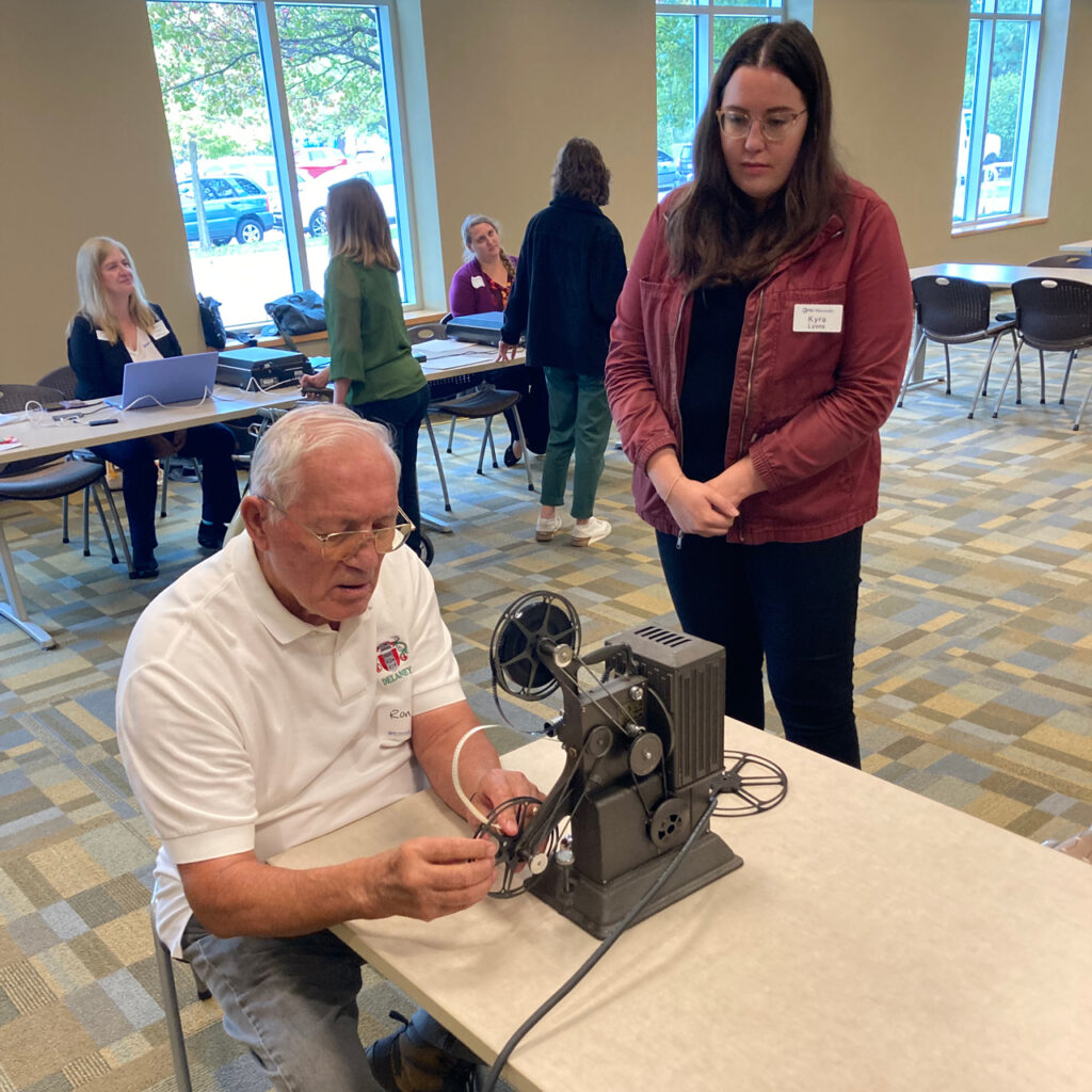 A man sets up an antique projector to play a movie for a young woman who is standing next to him as he sits at a table.