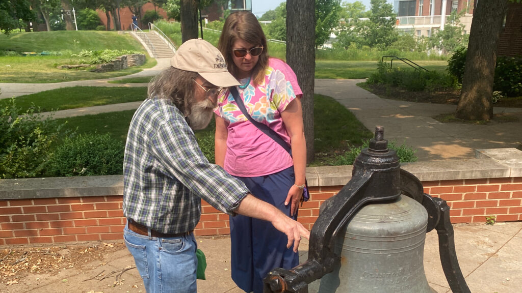 A man shows a woman an old tower bell that is on display at ground level on a college campus.