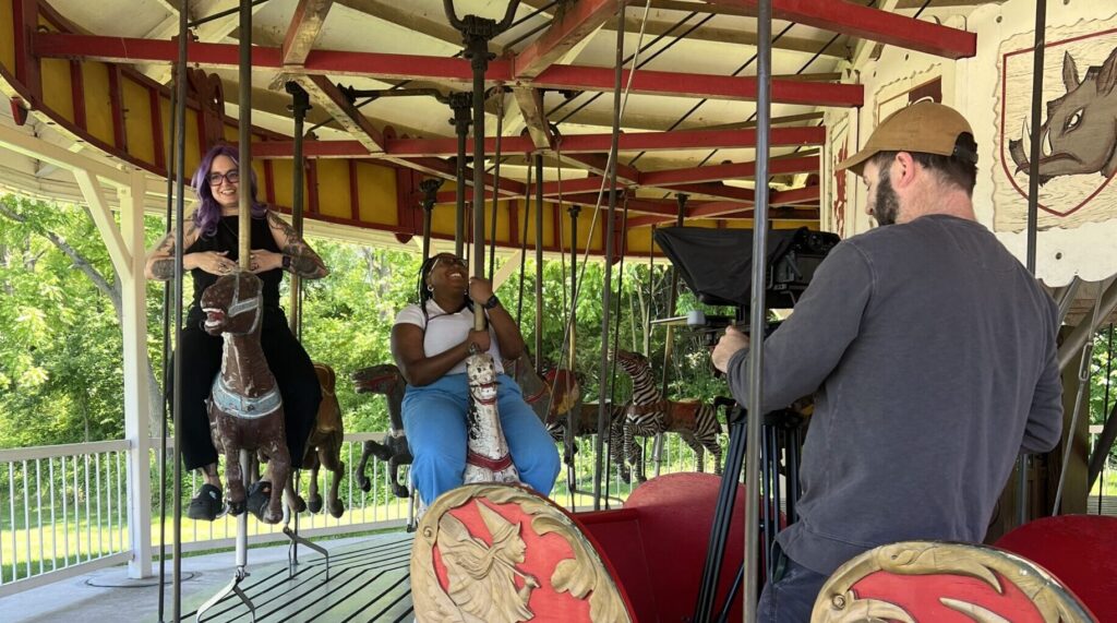 Two adults ride on a carousel while another adult films them.