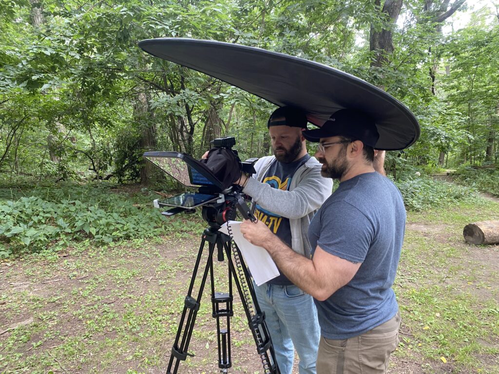 Two men stand outside in a forested area, looking at the display behind a camera.