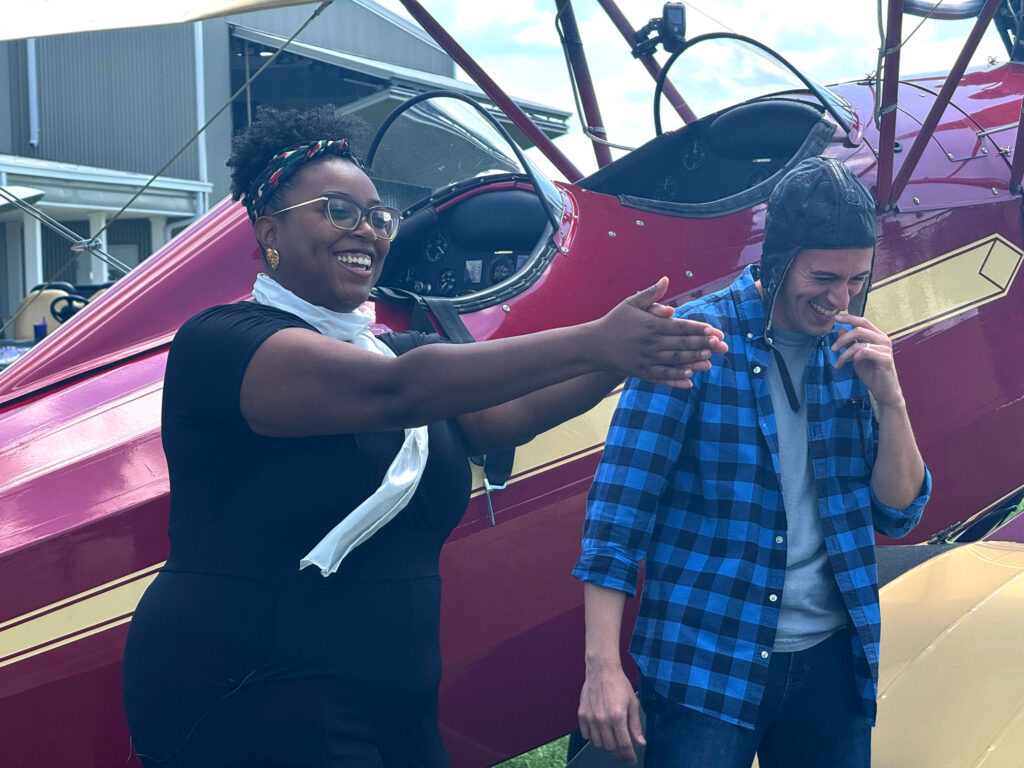 Two people stand in front of a red bi-plane.