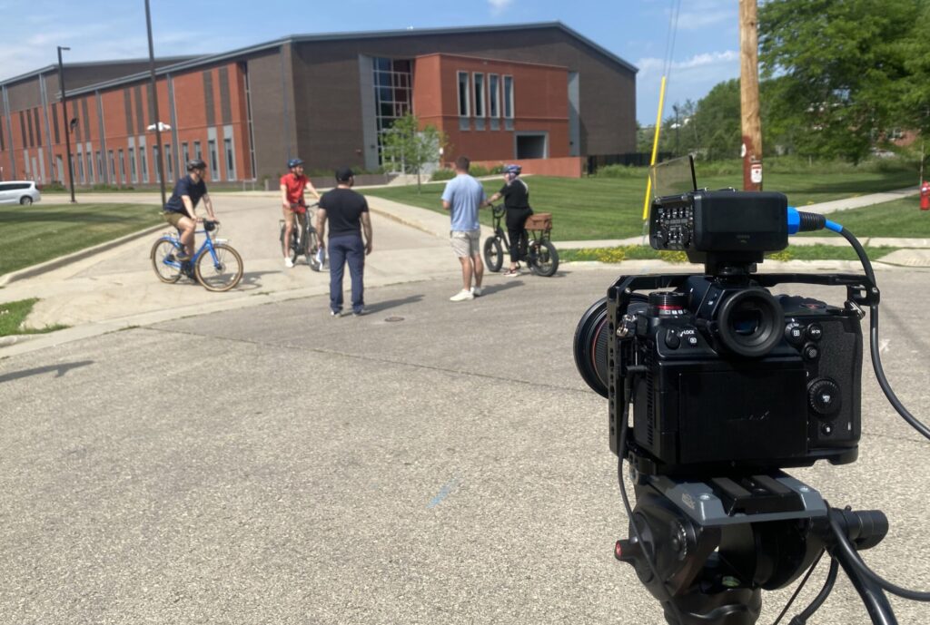 Cast and production crew film outside a red brick building. Three people are on bikes and wearing helmets.