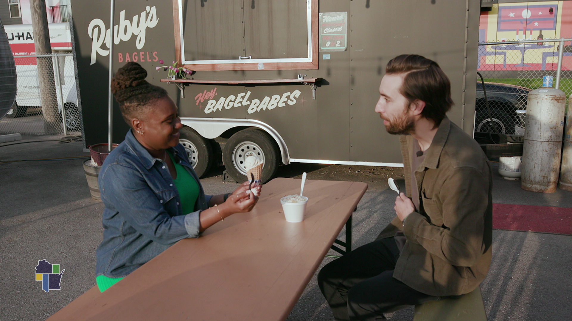 Wisconsin Life host Angela Fitzgerald eats ice cream with Zocálo Food Park co-owner Jesus Gonzalez on bench in front of Ruby's Bagels food truck at Zocalo Food Park.