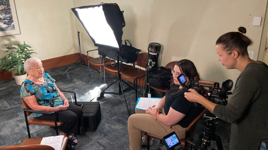 Two women set up lights and a camera to interview a woman sitting in a chair.