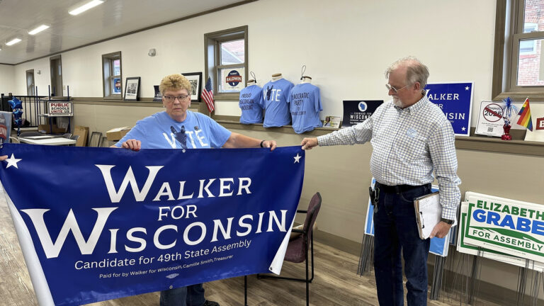 Joyce Bos stands and holds up a vinyl banner reading Walker for Wisconsin, Candidate for 49th State Assembly, and Paid for by Walker for Wisconsin, Camille Smith, Treasurer with a union label at bottom, while Scott Abbot Walker stands and holds one corner of the banner in his right hand, in a room with stacks of political yard signs leaning against a wall, torso mannequins wearing political t-shirts, other political signs and paraphernalia, and multiple chairs and tables.