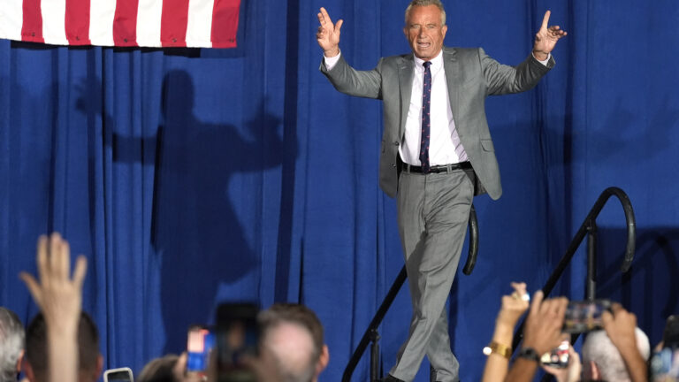 Robert F. Kennedy Jr. gestures with both hands in the air as he walks in front of hand railings on a stage, with his shadow on a stage curtain and the stripes of a U.S. flag in the background and out-of-focus raised hands and smart phones pointed toward him in the foreground.