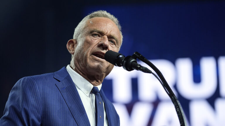 Robert F. Kennedy Jr. speaks into a microphone on a stand with an out-of-focus sign reading Trump Vance in the background.