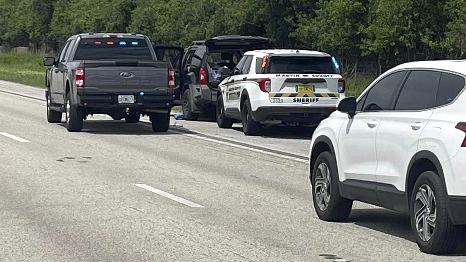 A police SUV and pickup truck park behind and next to another SUV on the shoulder of a multi-lane road as another SUV is stopped in the right lane in the foreground, with trees lining the road in the background.