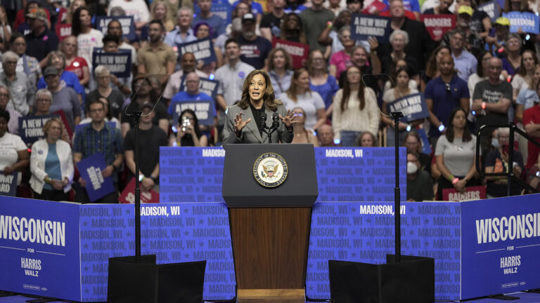 Kamala Harris gestures with both hands while standing and speaking into two microphones mounted to a wood podium with the Seal of the Vice President of the United States affixed to its front, with two teleprompter mirrors on either side and and a set of signs reading Wisconsin for Harris Walz and Madison, WI behind her, with out-of-focus people standing in the background, with some holding signs reading A New Way Forward and Badgers.