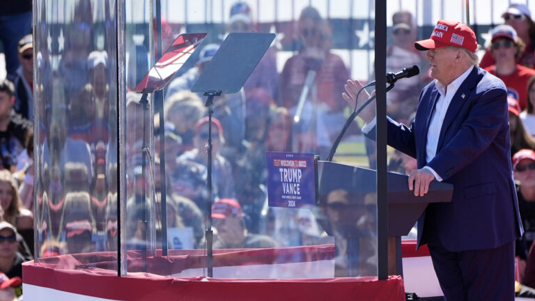 Donald Trump gestures with his right hand while speaking and standing at a podium and facing two teleprompter mirrors mounted behind a semicircular set of bullet-resistant glass panels, with out-of-focus seated people in the background.