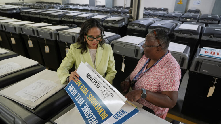 Paulina Gutierrez lifts a printed sign reading reading Welcome, Vote Here and Bienvenidos, Vote Aquí from a stack over multiple signs while standing next to Phyllis Whitley between two rows of ballot tabulator machines, with additional rows behind them in a large room with a structural support pillar.