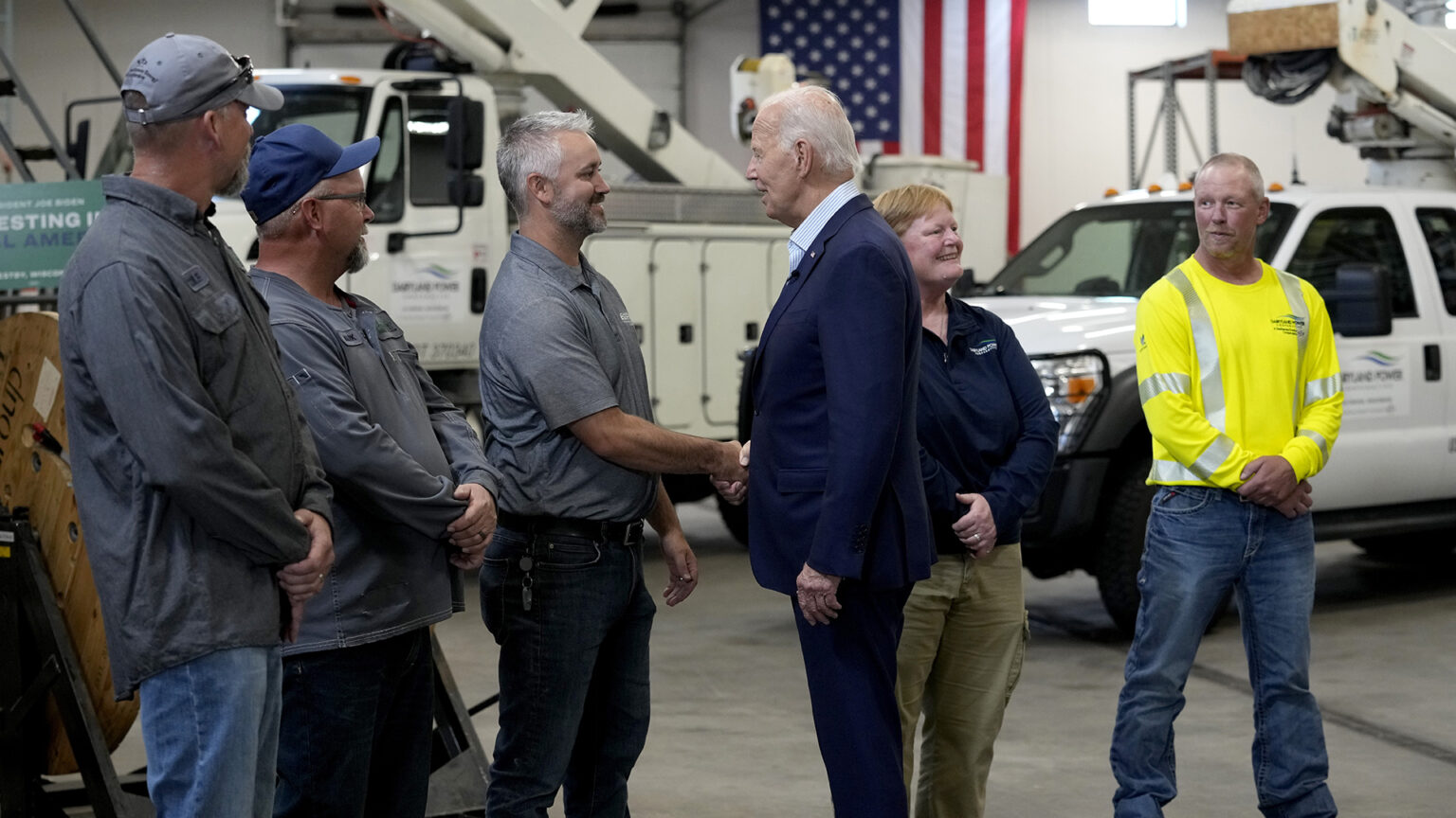 Joe Biden stands and shakes then hand of a person standing next to two others with their arms crossed in front of them, with two other people standing behind him, in a room with a concrete floor, parked utility vehicles and a U.S. flag on the back wall.