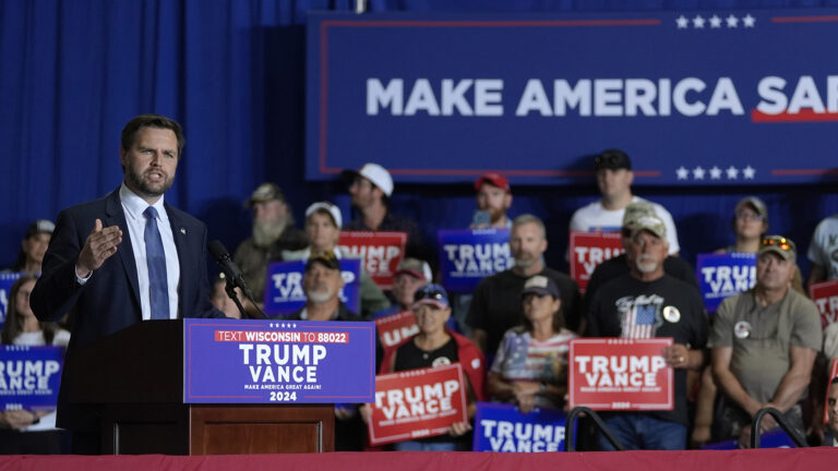 JD Vance speaks and gestures with his right hand while standing behind a podium with one mounted microphone and a Trump Vance 2024 campaign sign affixed to its front, with out-of-focus people standing behind him on risers while holding Trump Vance 2024 campaign signs in front of a stage curtain and large sign.