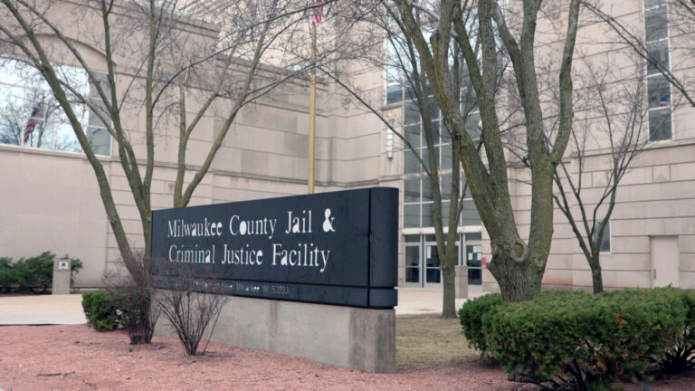 A large sign reading Milwaukee County Jail & Criminal Justice Facility with an address stands amid bushes and trees in front of a masonry and glass, with multiple glass doors on one wall.