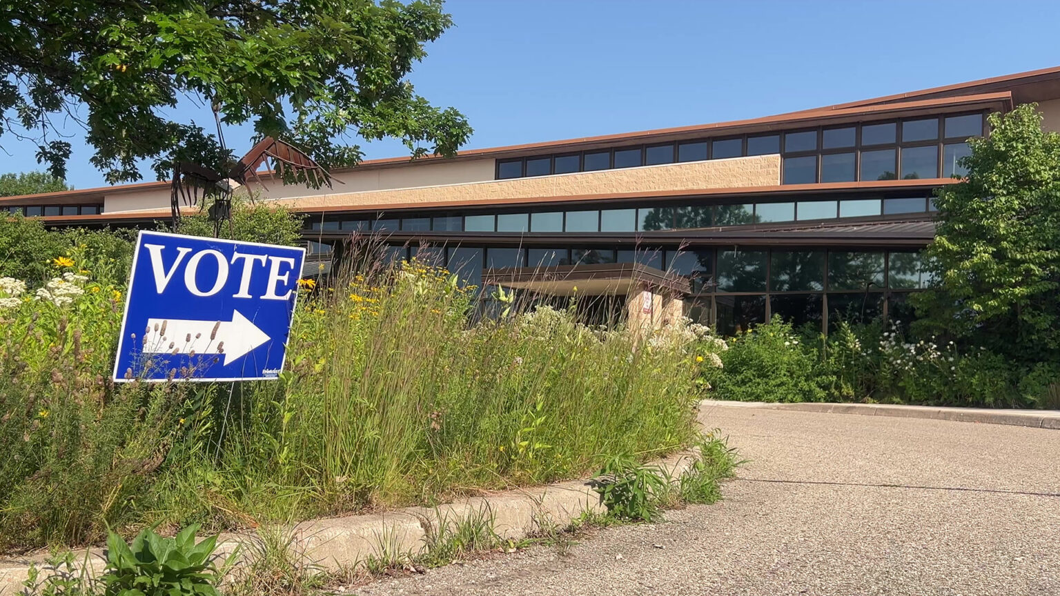 A political yard sign with the word VOTE and an arrow is placed amid high prairie grasses in the median of a roundabout, with a masonry and glass Prairie Style-building and trees in the background.