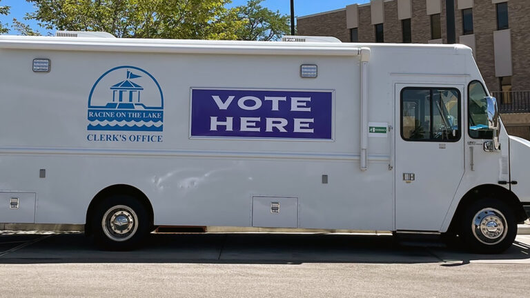 A step van with the logo of the City of Racine Clerk's office logo and the words VOTE HERE painted on its side is parked in a parking lot, with trees and a building in the background.