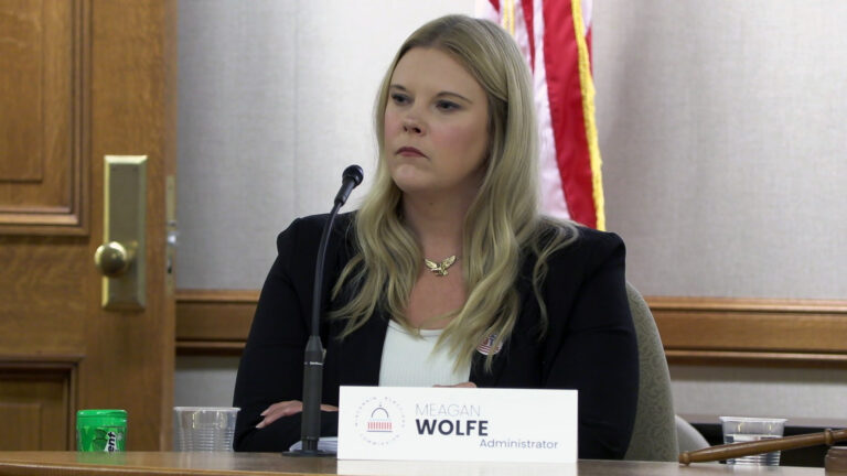 Meagan Wolfe crosses her arms in front of her while sitting at a table with a mounted microphone, plastic cups and a paper sign reading Meagan Wolfe and Administrator, in a room with a wood door with a brass doorknob and a U.S. flag in the background.