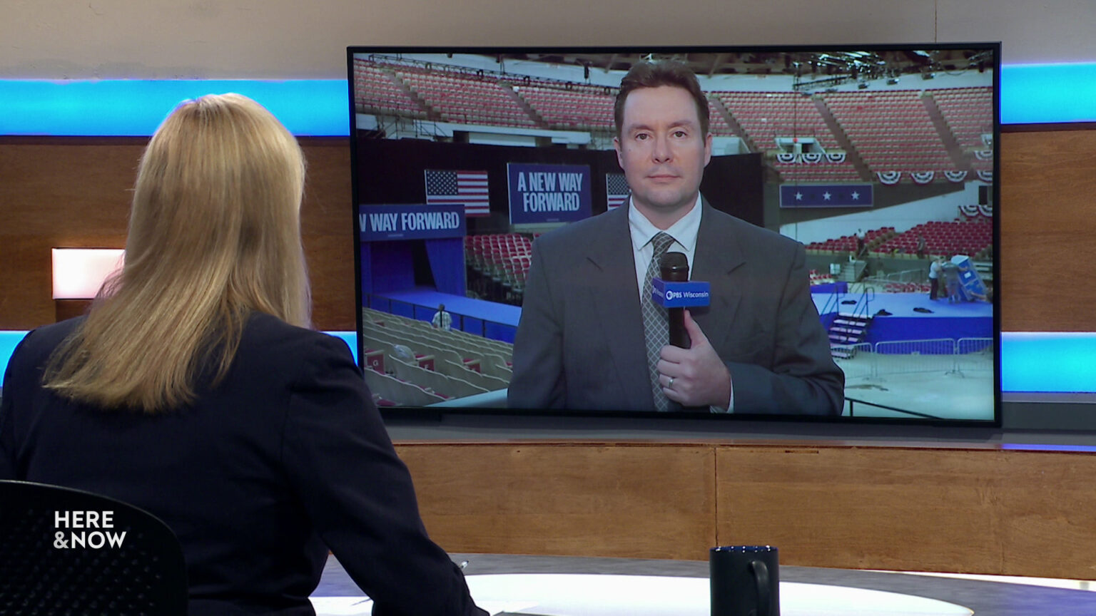 Frederica Freyberg sits at a desk on the Here & Now set and faces a video monitor showing an image of Zac Schultz.