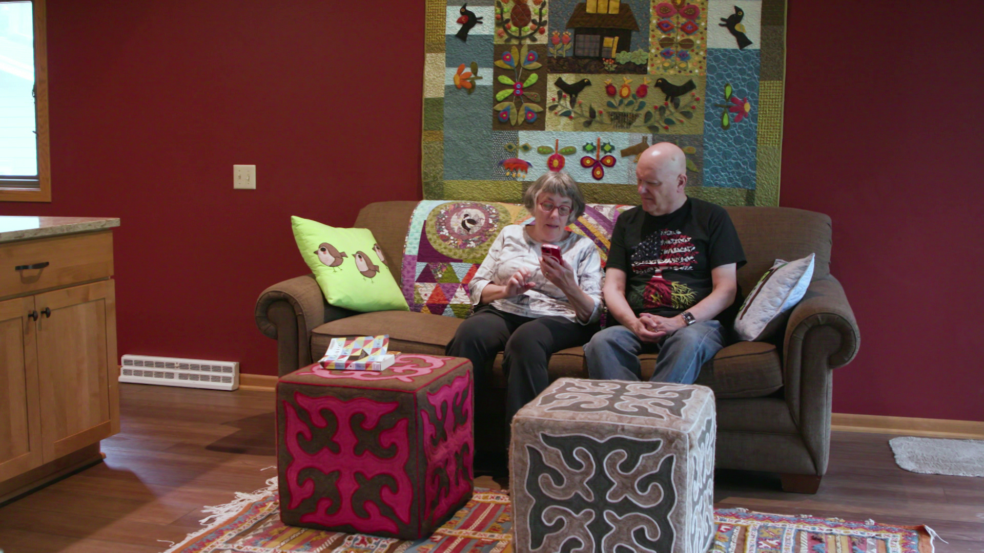 Toni and Mark Swandby sit on a couch and look at a phone ina room with two square ottomans on a rug with a quilt on the wall behind them and a countertop to the side.