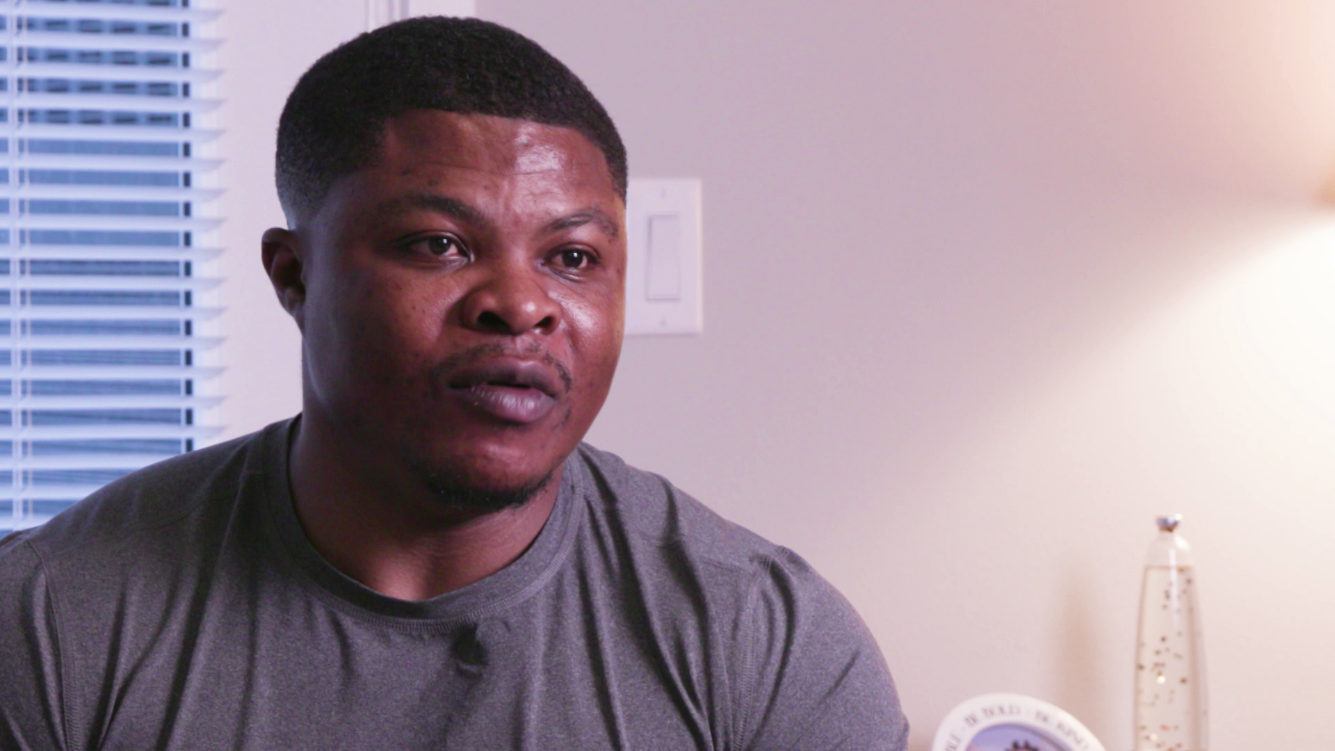 Ngwa Augustine sits while speaking indoors with a window with blinds and light switch in the background.