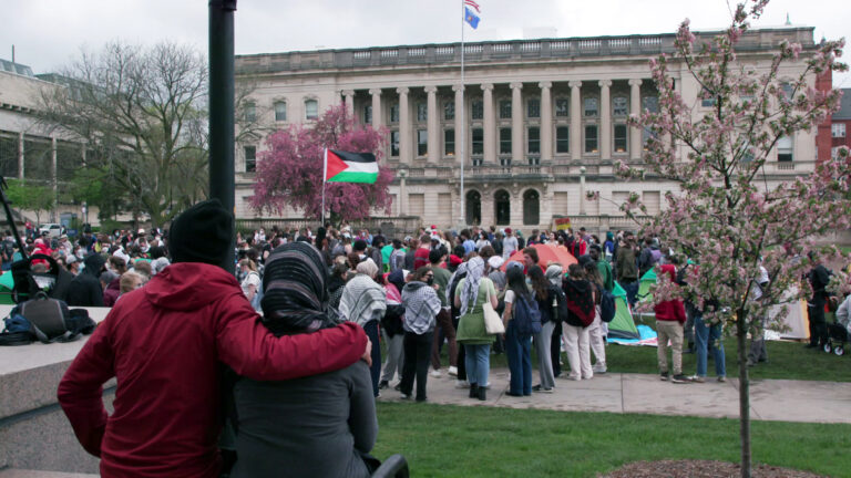 Two people stand next to a blossoming tree and face a larger group of protestors, some wearing kaffiyehs and one holding a Palestine flag on a hand-held pole, stand on sidewalks and lawns around multiple personal camping tents outdoor area, with a flagpole with the US. and Wisconsin flags in front of a masonry building with balustrades and an adjacent concrete Brutalist-style building in the background.