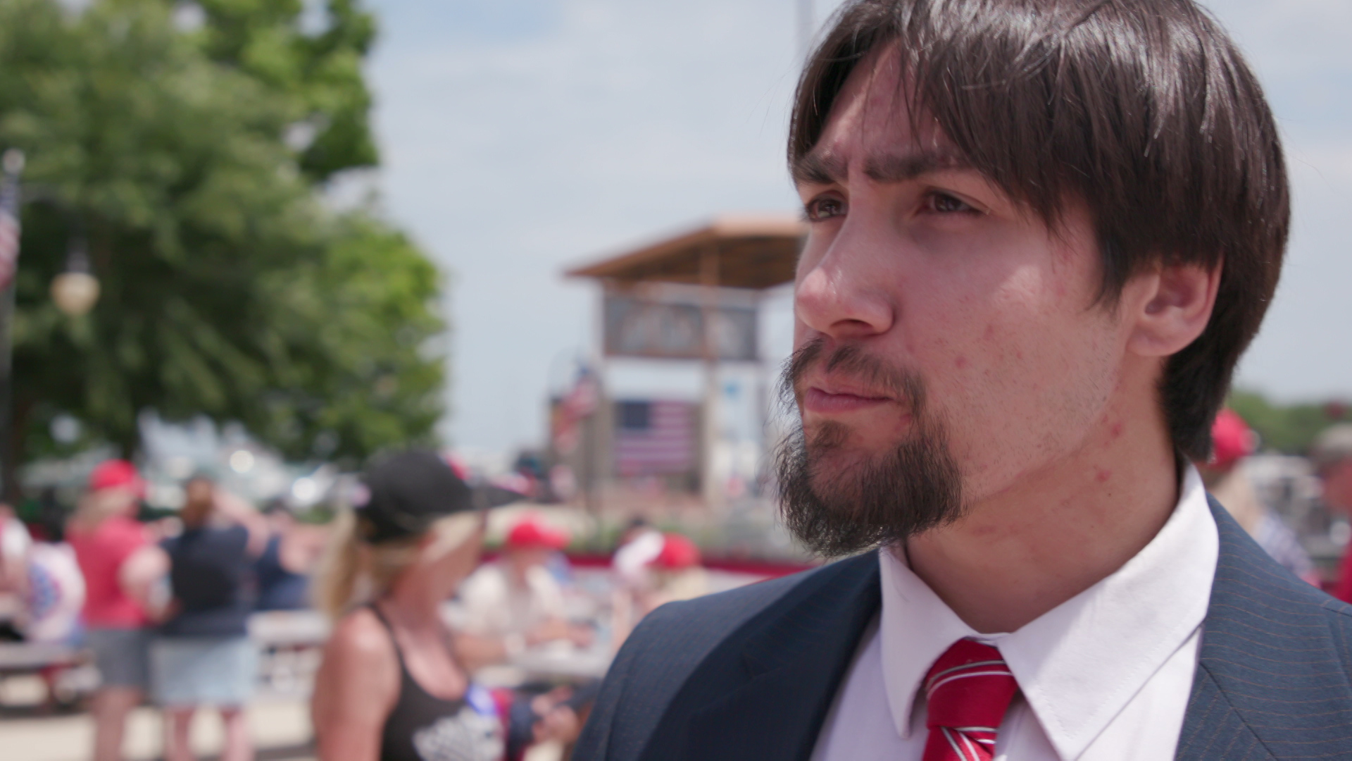 Hilario Deleon speaks while standing outside with out of focus people walking, trees and a stage in the background.