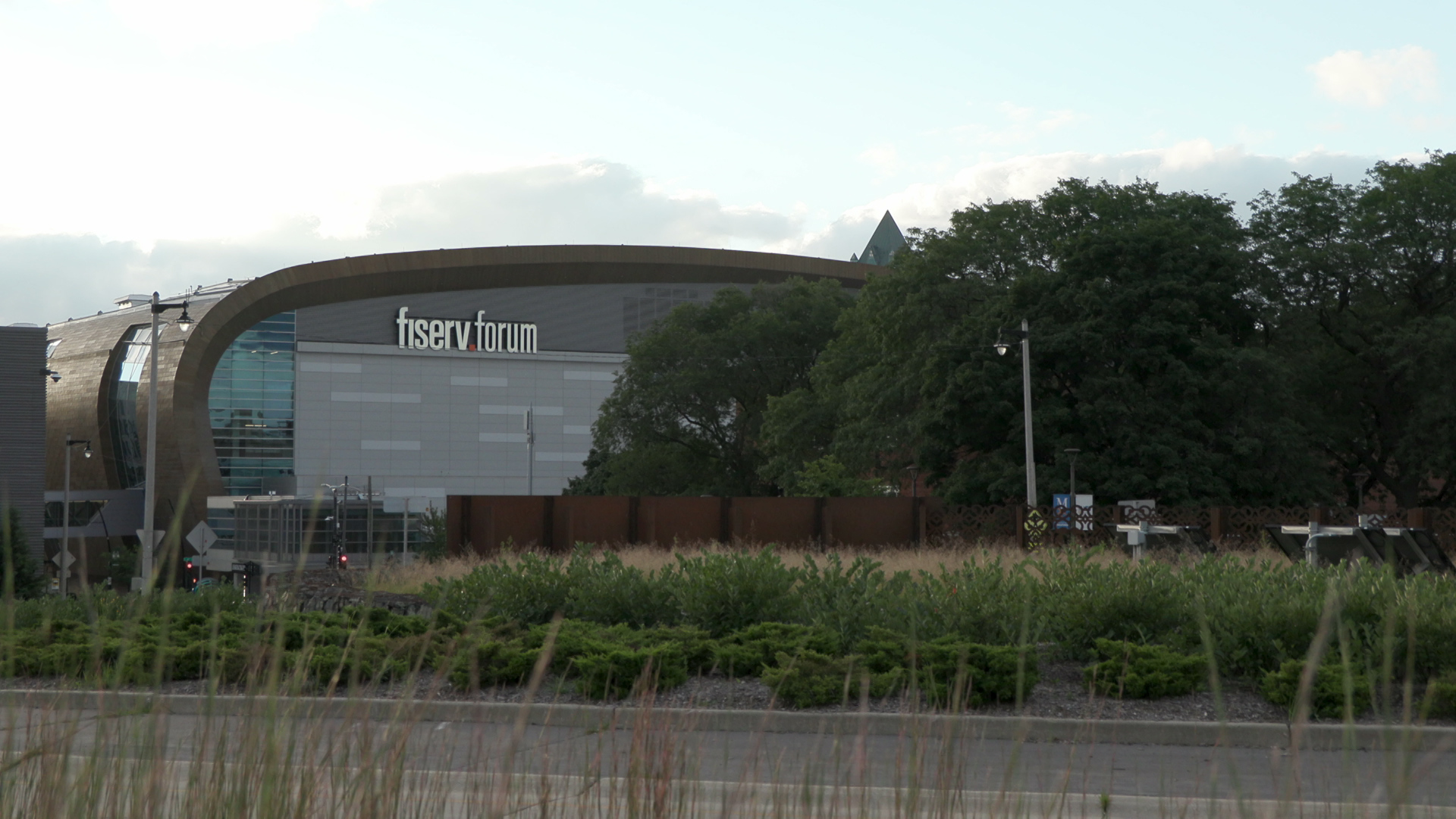 A building with words reading "fiserv.forum" on one side stands behind a group of trees with fencing, bushes and a road in the foreground.