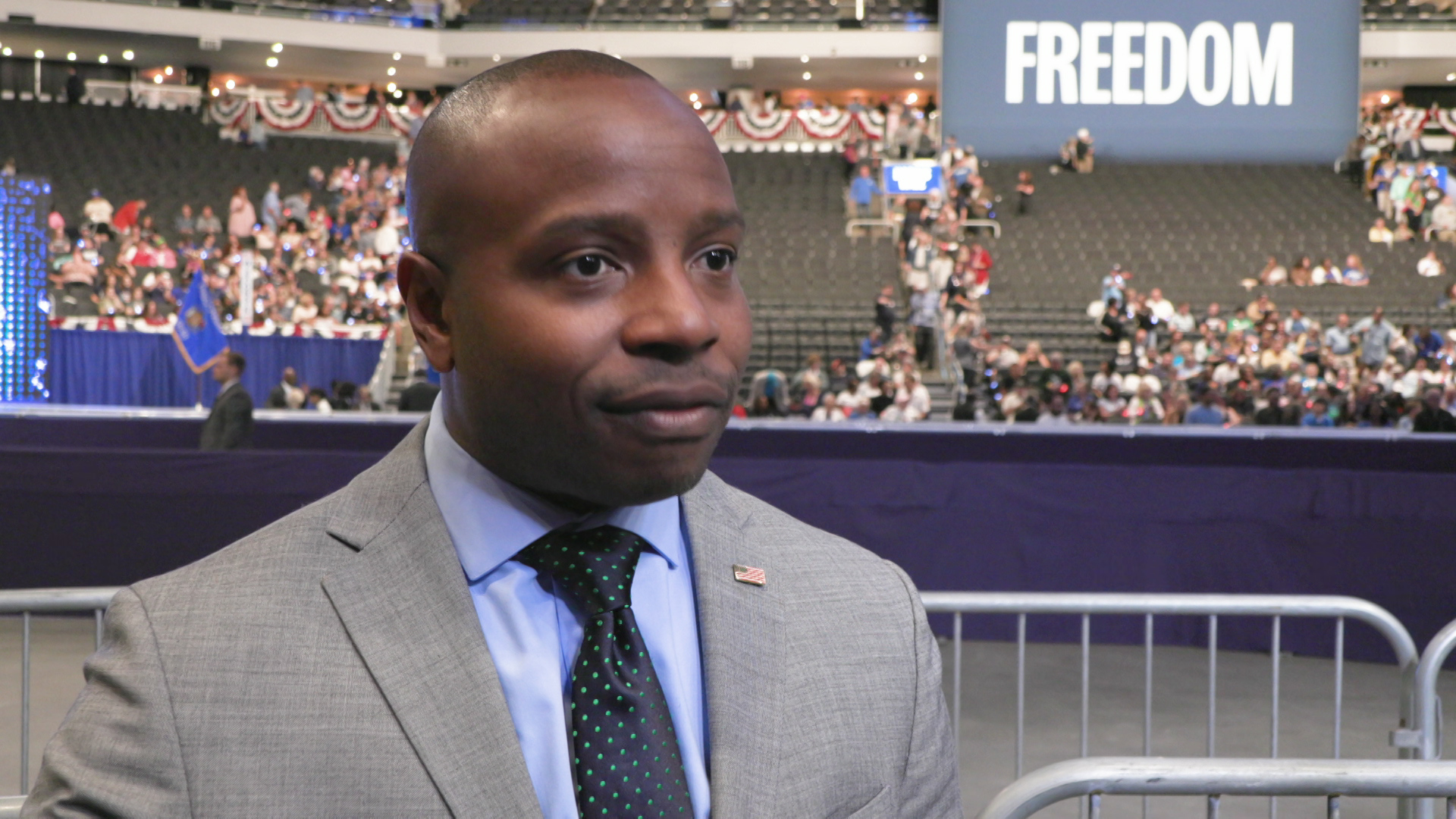 Cavalier Johnson speaks while standing on the floor of an arena with out of focus people sitting in ascending rows of seats, with red, white and blue bunting displayed on railings next to an electronic sign reading "Freedom."