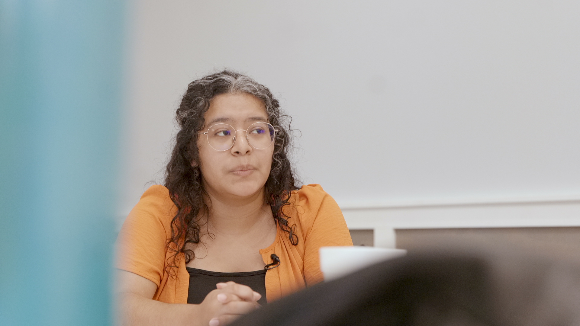 Priscilla Perez speaks while siting with her hands folded in front her with out of focus objects in the foreground.