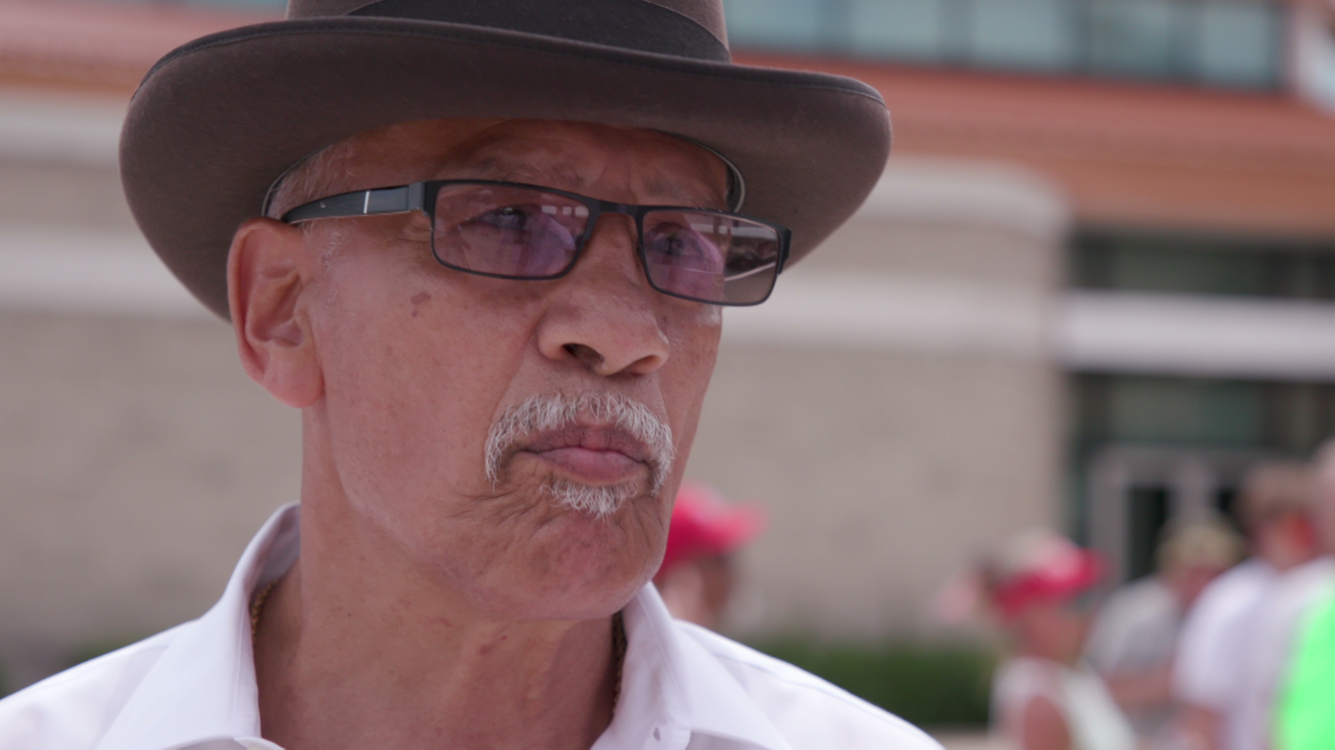 Antonio Servantez speaks while standing outside with out of focus people walking and a building in the background.