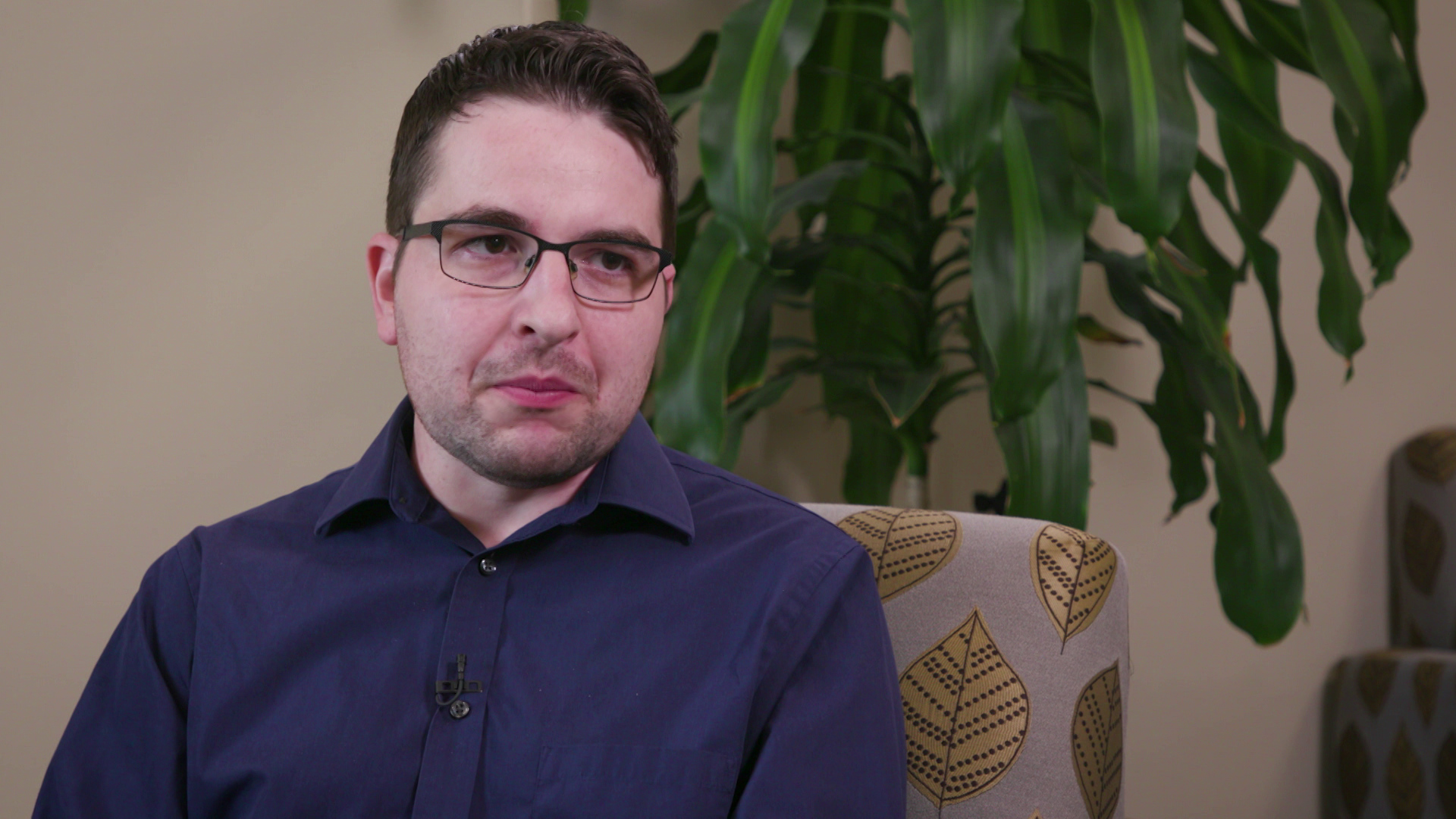 Anthony Chergosky speaks while sitting in a chair with a leaf design in a room with a potted plant in the background.