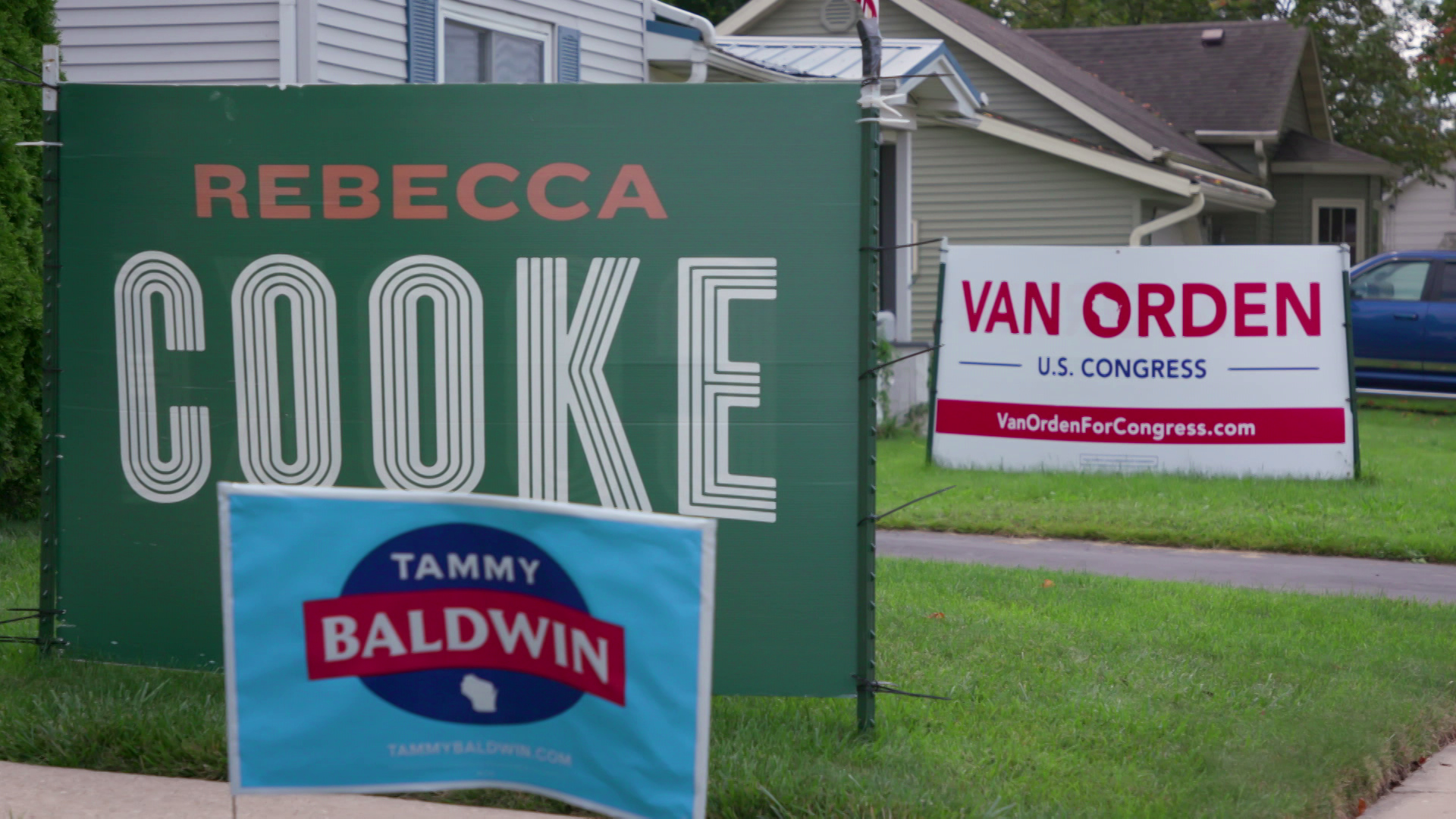 Political signs reading "Van Orden U.S. Congress," "Rebecca Cooke" and "Tammy Baldwin" sit on patches of grass in between sidewalks with houses and a car in the background.