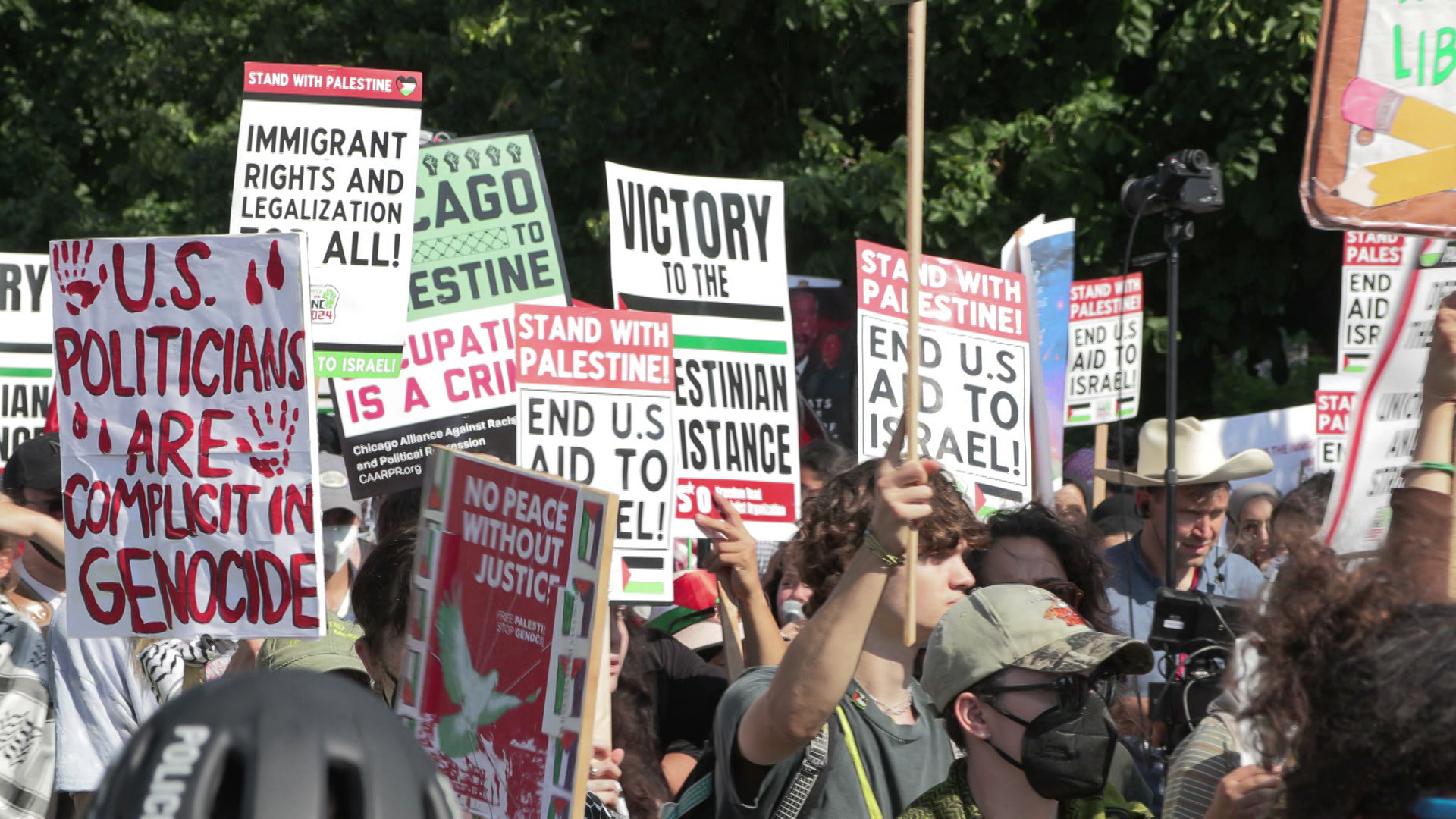 People march with multiple signs related to the Israel-Hamas conflict while outside with trees in the background.